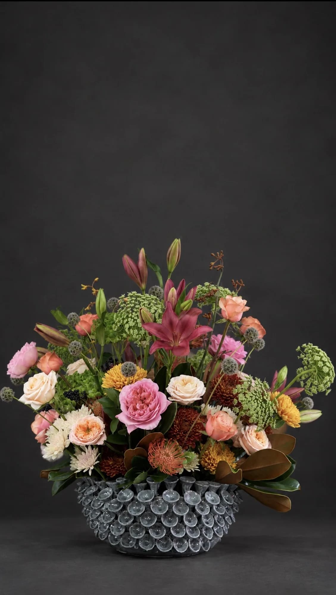 Garden-style arrangement of pink and peach roses, red lilies and protea in a scalloped grey vessel