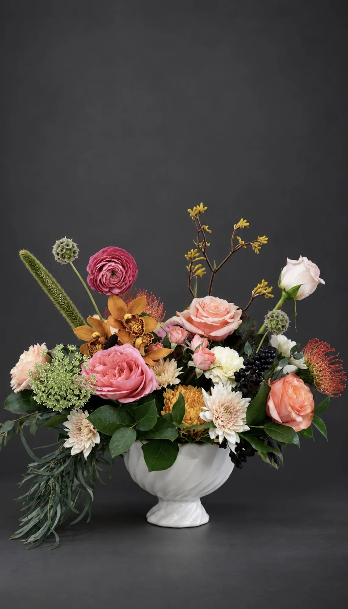 Pink roses, ranunculus and protea with trailing greenery in a white footed compote